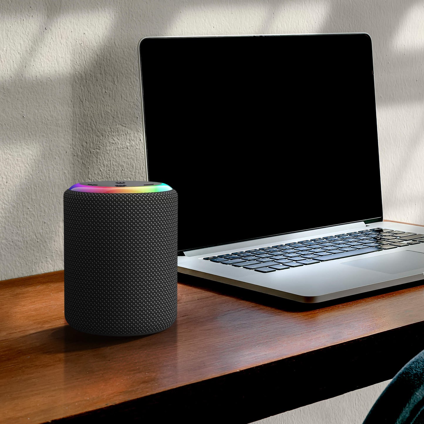 Photograph of a black textured smart speaker with a rainbow RGB light ring next to an open silver laptop with a blank screen on a wooden desk